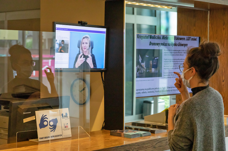 Lada przy kasie Muzeum MOCAK. Za szybą plakietka z piktogramem tłumacza języka migowego. Przed ladą stoi kobieta, miga. Nad wysokości jej wzroku zawieszony ekran z kamerą, na ekranie tłumacz języka migowego.