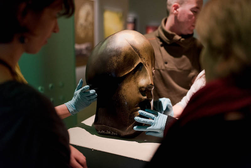 Na podeście ustawiona rzeźba przypominająca ludzką głowę o nieregularnym kształcie. Dookoła kilka osób w lateksowych rękawiczkach, dotykają rzeźby z różnych stron.