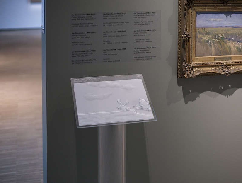 Fragment ekspozycji Muzeum Śląskiego. Pod ścianą z obrazami ustawiona tyflografika, na przezroczystym podeście, ustawiona pod kątem 45 stopni, tyflografika przedstawia jeden z obrazów na wystawie.