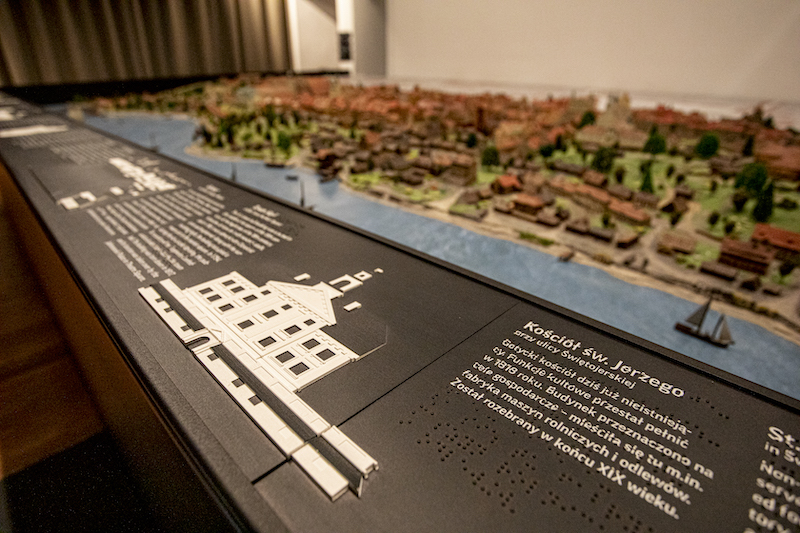 Fragment makiety. Przy krawędzi czarny panel z elementami dotykowymi. Wypukły kształt elewacji kościoła, obok treść opisu w druku i w Braille’u.