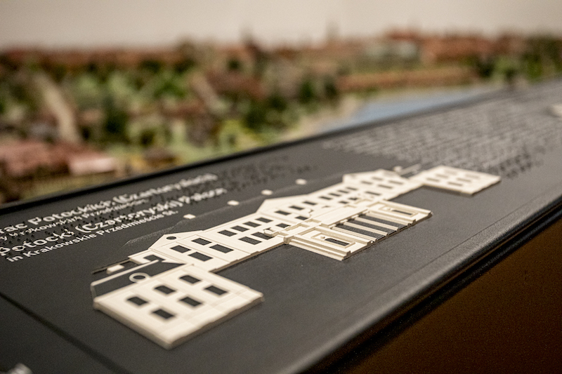 Fragment makiety. Przy krawędzi czarny panel z elementami dotykowymi. Wypukły kształt elewacji pałacu Potockich, obok treść opisu w druku i w Braille’u.