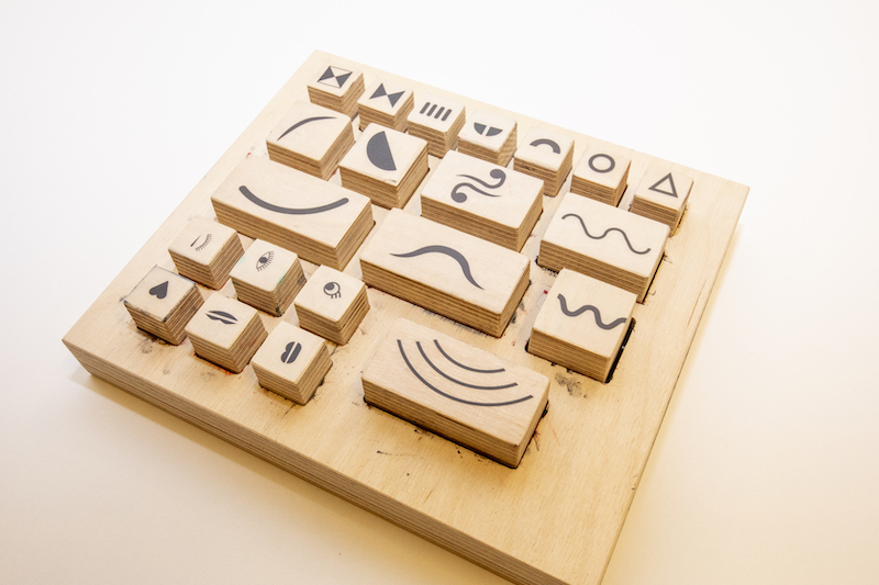 Drewniana plansza z wypukłymi, ruchomymi kostkami. Na każdej z nich narysowany kształt lub symbol.