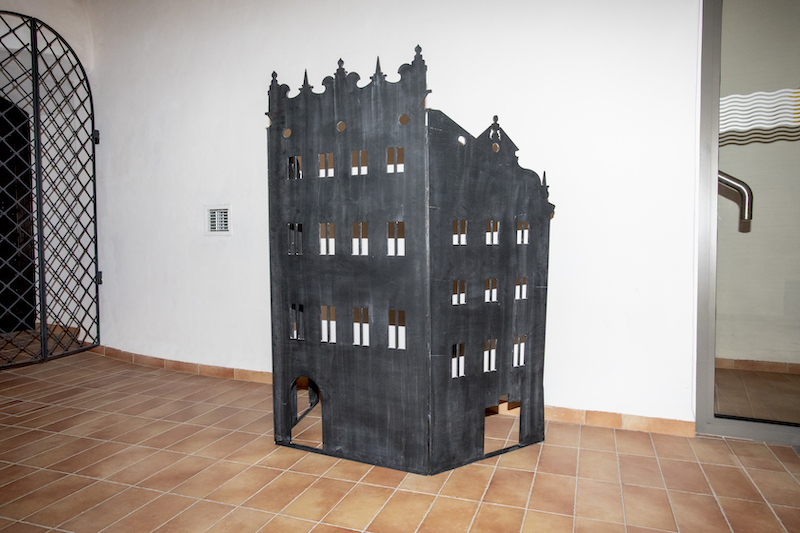 Kartonowy model frontu ozdobnej kamienicy. Całość pomalowana na czarno, w materiale wycięte są drzwi i okna, a także zdobienia na szczycie budynku.