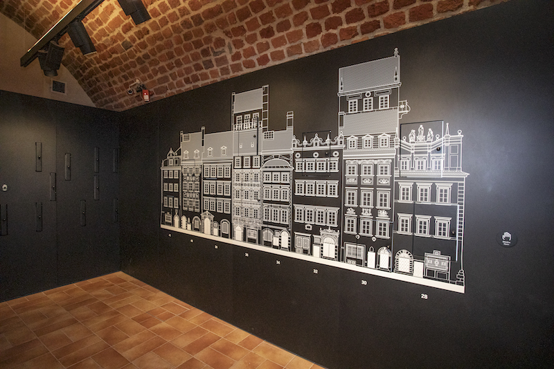 Pomalowana na czarno ściana. Na niej białe wypukłe elementy obrazujące elewacje kamienic Starego Miasta. Zaznaczone są okna, drzwi, dachy, zdobienia. W ścianie są ruchowe schowki i szuflady.