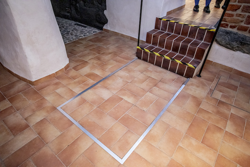 Korytarz. Trzy stopnie prowadzące z jednego poziomu na drugi. Przy stopniach na niższym poziomie kształt prostokąta zaznaczony w podłodze metalową listwą.