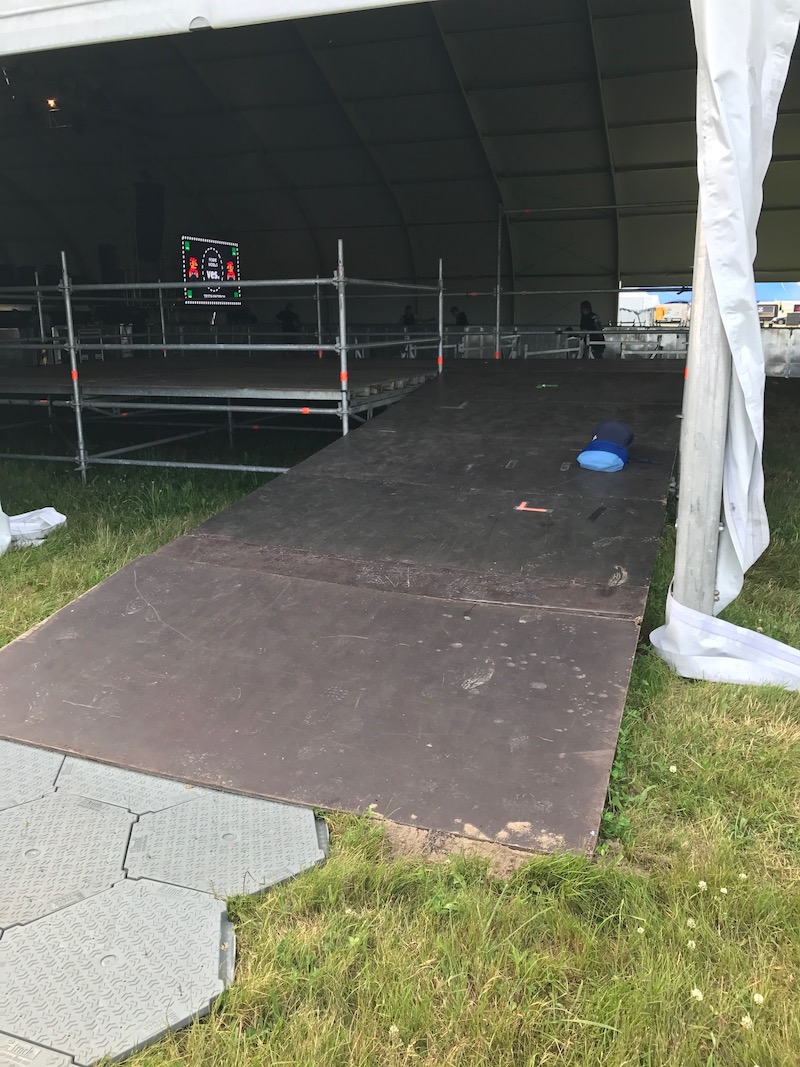 Zdjęcie z festiwalu plenerowego. Ogromny namiot koncertowy, w nim ogrodzony podest. Do środka prowadzą rozłożone na trawie maty i szeroka pochylnia.