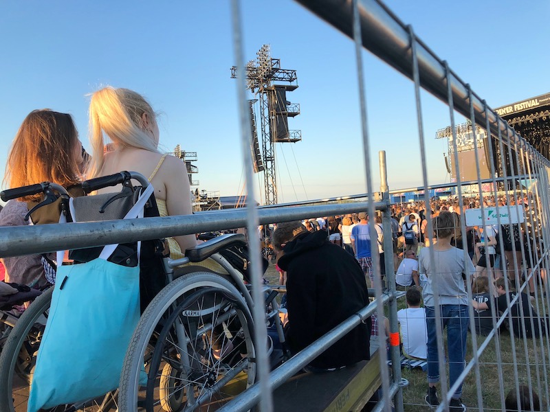 Festiwal plenerowy. Na podwyższonej ogrodzonej platformie kilka osób na wózkach. Z poziomu platformy jest ułatwiony widok na scenę, ponad głowami pozostałych uczestników.