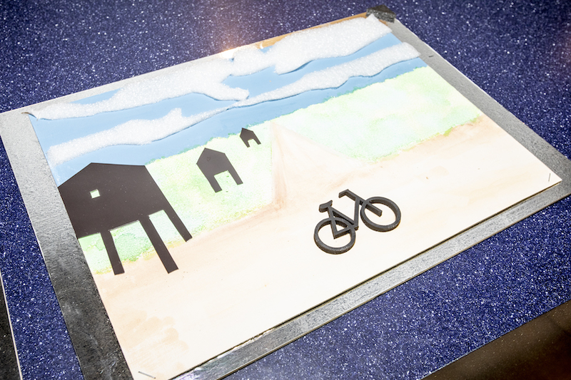Tyflografika obrazu przedstawiającego pejzaż, wykonana z materiałów o różnych fakturach. Chmury z gabki, droga z ziarnistej farby, budynki z naklejonego grubego kartonu, kształt roweru wycięty z plastiku.