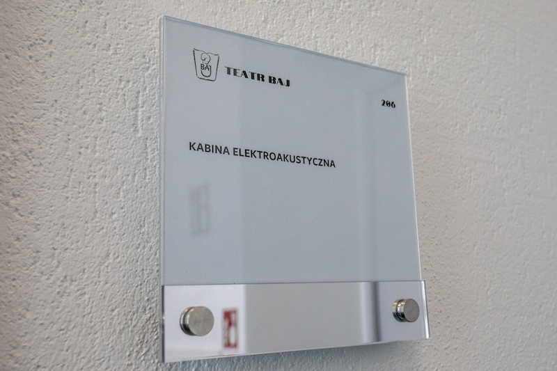 Ściana przy drzwiach. Na białej szklanej tabliczce logotyp Teatru Baj oraz napis kabina elektroakustyczna.
