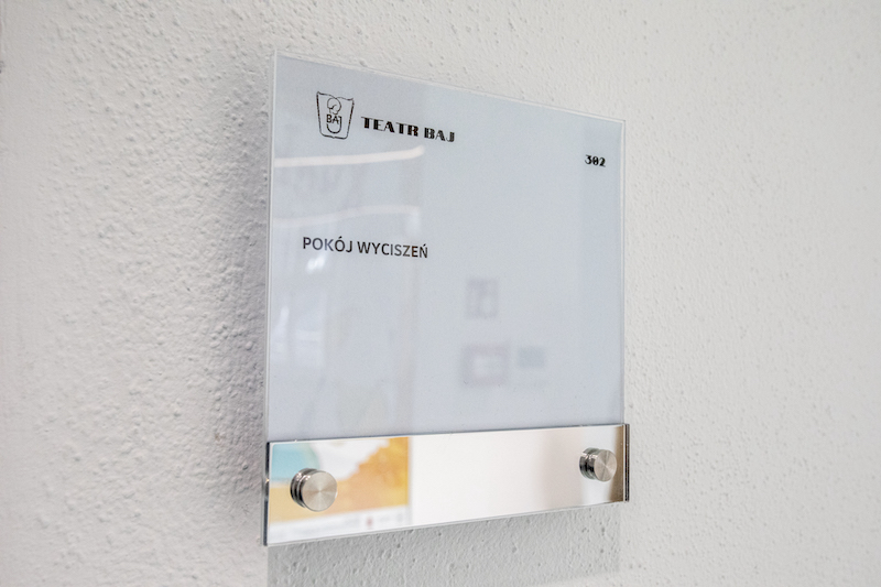 Ściana przy drzwiach. Na białej szklanej tabliczce logotyp Teatru Baj oraz napis pokój wyciszeń.