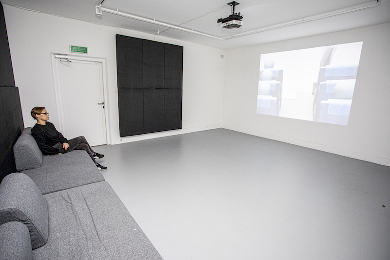 Sala wystawowa. Na ścianie wyświetlony film z rzutnika. Przy przeciwległej ścianie ustawiony rząd kanap. Na jednej z nich siedzi osoba, ogląda wyświetlany film.