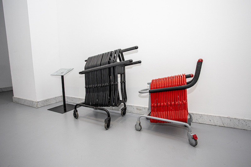 Korytarz przy wejściu na wystawę. Dwa wózki, na których zawieszone są przenośne składane taborety. Na większym wózku większe czarne taborety, na mniejszym - małe czerwone.