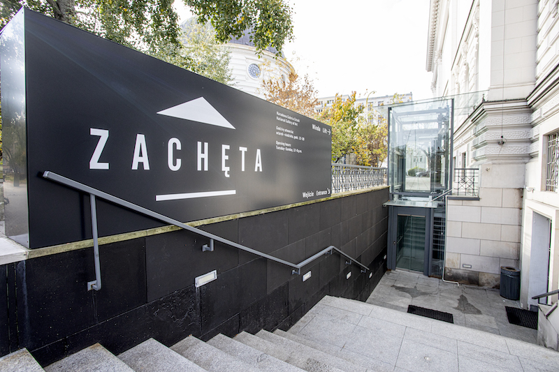 Wejście boczne do budynku Galerii Zachęta. Drzwi znajdują się niżej niż poziom chodnika. Do nich prowadzą schody z poręczą, a z przeciwnej strony przeszklona winda.